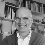 A well-dressed person with glasses smiles in front of a bookshelf filled with various books.